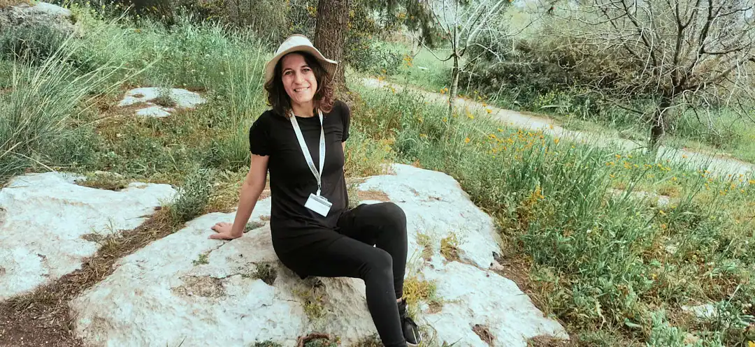 נטע מטיילת יושבת על סלע בשטח פתוח, בין עשבים ואבנים